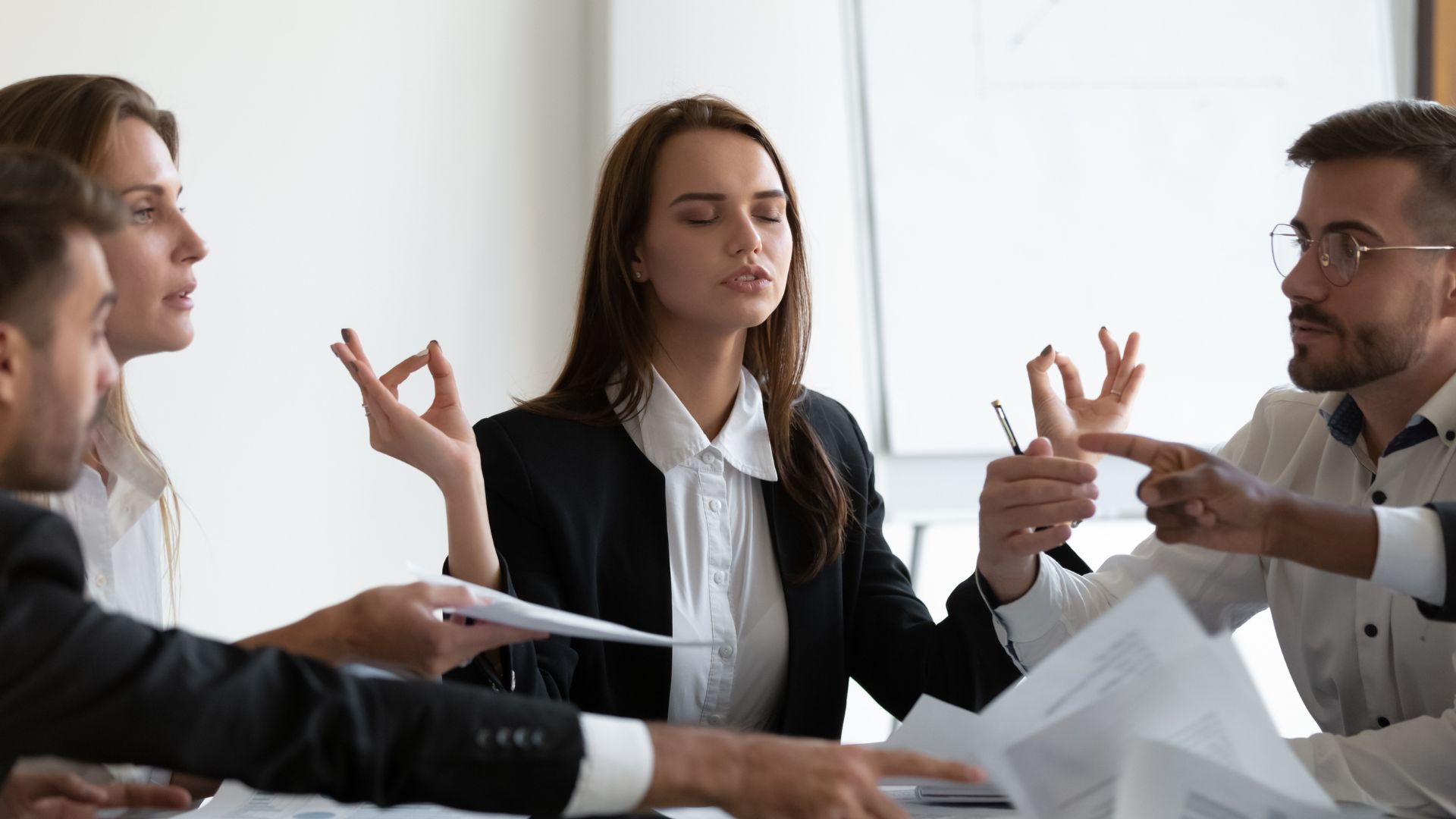 Mindful young business woman meditating managing stress, ignoring annoyed arguing colleagues at workplace.